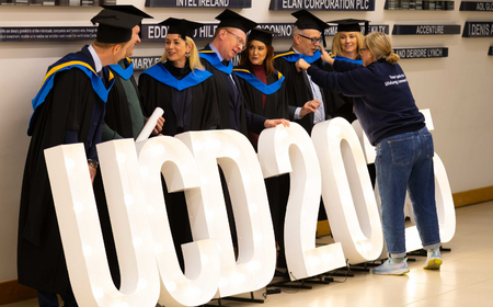 UCD graduates in robes posing with large illuminated “UCD 2025” letters.